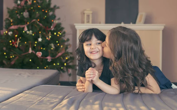 Elder sister kiss the younger. Girls sitting on the floor near a Christmas tree and presents. — Stock Photo, Image