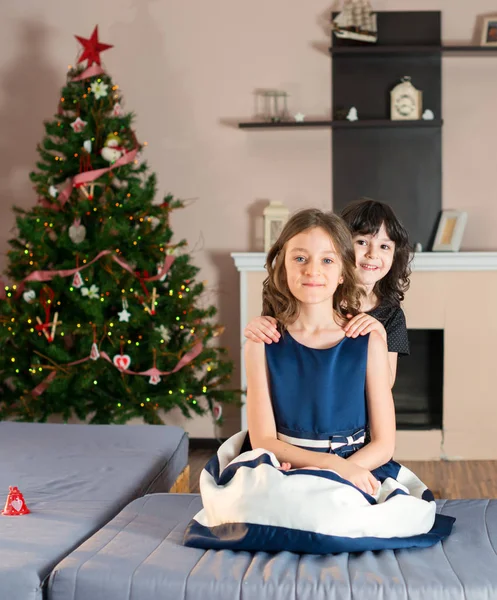 Elder sister with the younger one. Girls sitting on the couch near a Christmas tree and presents. — Stock Photo, Image
