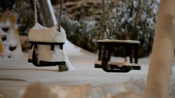 Balançoire solitaire se balançant d'un côté à l'autre dans un parc d'hiver abandonné couvert de neige éclairée par la lumière du lampadaire . — Video