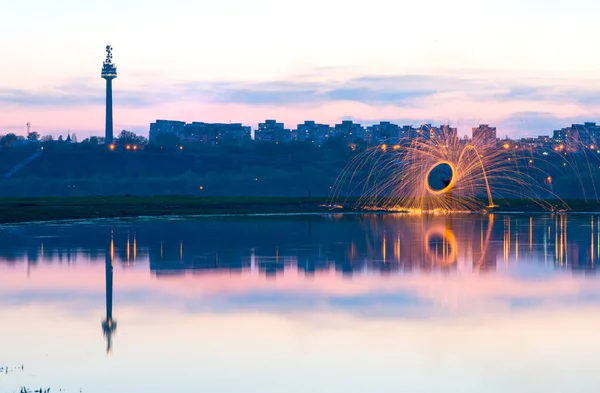 Hot Golden Sparks Flying from Man Spinning Burning Steel Wool near River with Water Reflection. Long Exposure Photography using Steel Wool Burning