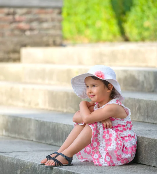 Adorable niña pequeña con sombrero blanco sentado en las escaleras en el día de verano cálido y soleado —  Fotos de Stock