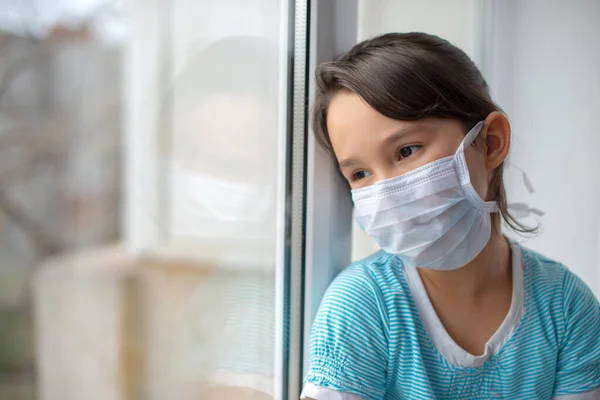可爱的伤心的小女孩戴着医疗面罩站在家里的窗边 Coronavirus大流行病 — 图库照片