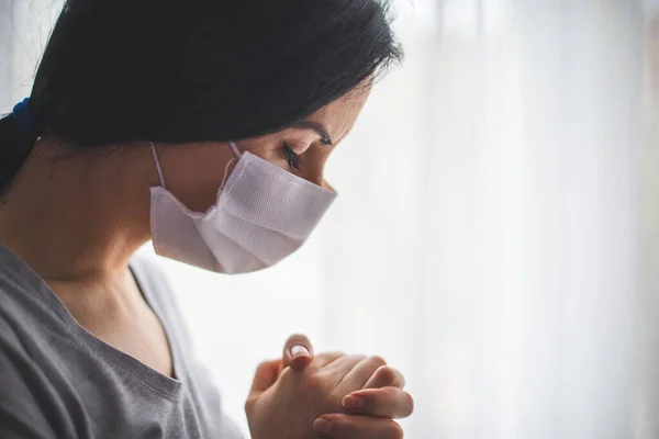 窓の横で祈っている外科的マスクを持つ女性の肖像 — ストック写真