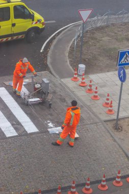 Trafik çizgisi boyama. İşçiler yaya geçidinde beyaz sokak çizgileri çiziyorlar. Arka planda turuncu ve beyaz çizgili yol konileri, ayakta