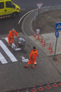 Trafik çizgisi boyama. İşçiler yaya geçidinde beyaz sokak çizgileri çiziyorlar. Arka planda turuncu ve beyaz çizgili yol konileri, ayakta