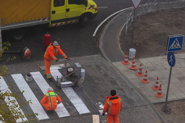 Traffic line painting. Workers are painting white street lines on pedestrian crossing. Road cones with orange and white stripes in background, standing