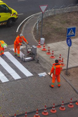 Trafik çizgisi boyama. İşçiler yaya geçidinde beyaz sokak çizgileri çiziyorlar. Arka planda turuncu ve beyaz çizgili yol konileri, ayakta