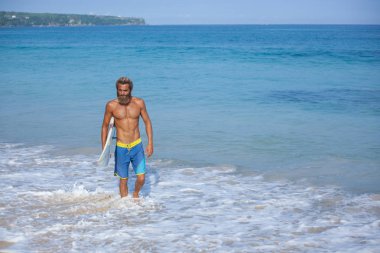 Bir surfboard ile çekici sakallı kıvırcık adam çerçevenin sol tarafında bulunan bir plajda yürüyor