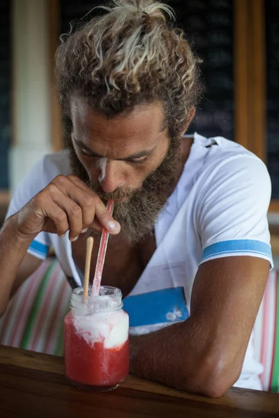 Привлекательный бородатый кудрявый мужчина с кружкой смузи сидит в кафе и пьет через соломинку — стоковое фото