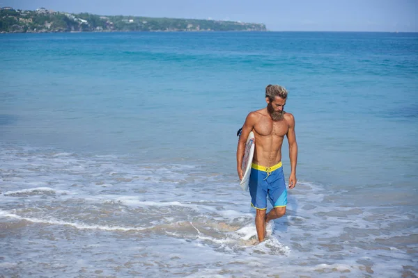 Привлекательный бородатый кудрявый мужчина с доской для серфинга ходит по пляжу, расположенному на правой стороне рамы — стоковое фото