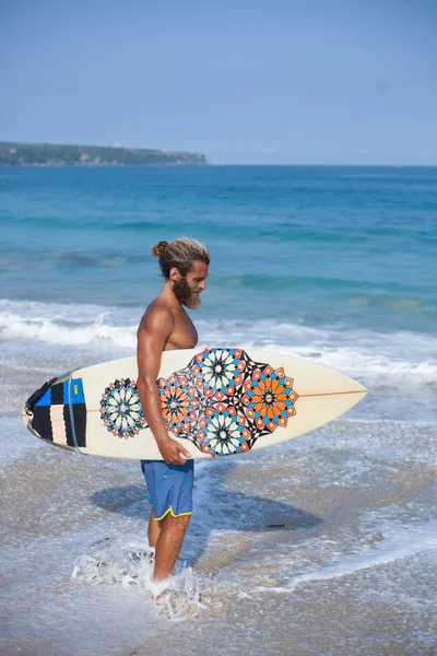 Das vertikale Porträt eines attraktiven, bärtigen, lockigen Mannes mit Surfbrett steht an einem Strand — Stockfoto