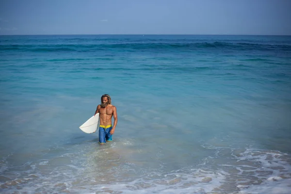 Atraktivní vousatých kudrnaté mokrý muž v modrých kalhotách a s nahý trup s bílým Surf přichází z oceánu — Stock fotografie