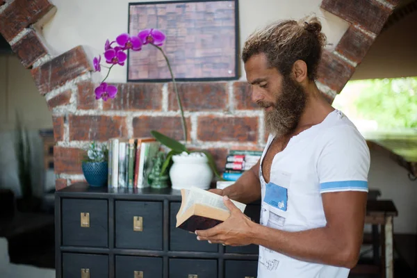 Портрет модного бородатого человека, стоящего возле книжной полки в кафе и читающего книгу — стоковое фото