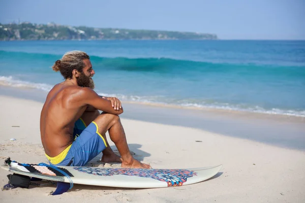 Attraktiver bärtiger Lockenkopf mit Surfbrett sitzt mit dem Rücken auf einem Sand und blickt auf den Ozean — Stockfoto