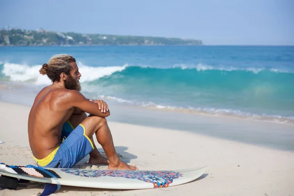 Attraktiver bärtiger Lockenkopf mit Surfbrett sitzt mit dem Rücken auf einem Sand und blickt auf den Ozean — Stockfoto