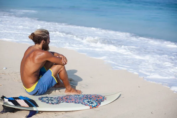 Attractive bearded curly man with a surfboard is seating with his back on a sand and looking to the ocean