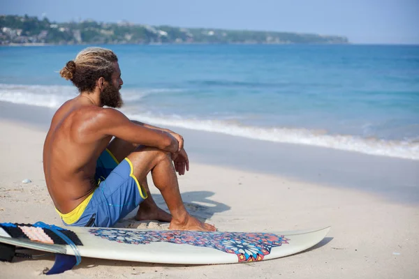 Porträt eines attraktiven bärtigen lockigen Mannes mit Surfbrett sitzt mit dem Rücken auf einem Sand und blickt auf den Ozean — Stockfoto