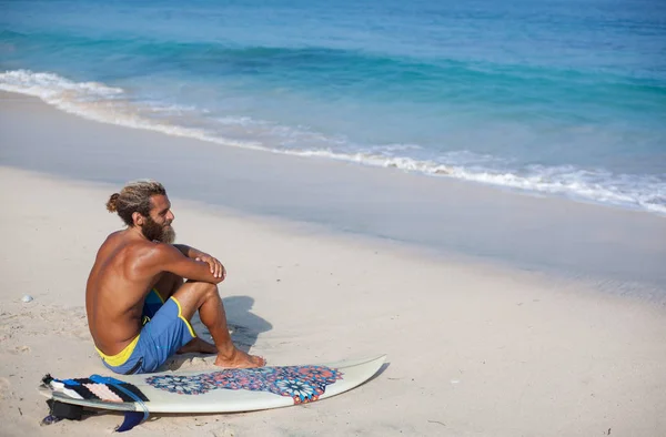 Porträt eines attraktiven bärtigen lockigen Mannes mit Surfbrett sitzt mit dem Rücken auf einem Sand und blickt auf den Ozean — Stockfoto