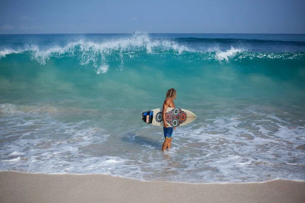 Kudrnaté blond aristokratickým krasavcem surfovací prkno, stojící na břehu moře a při pohledu na velké vlny, za ním — Stock fotografie