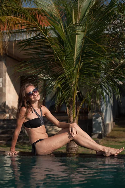 Retrato vertical de una hermosa chica en bikini negro y gafas de sol sentadas en el borde de la piscina, una casa y una palmera en el fondo — Foto de Stock
