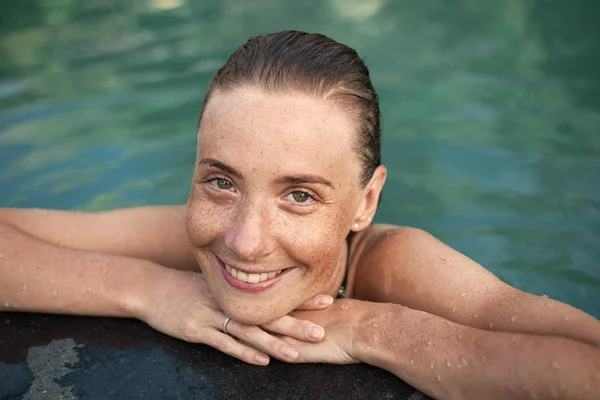 Retrato horizontal de cerca de una hermosa chica sonriente con pecas y cabello mojado en el agua, brazos en el borde de la piscina —  Fotos de Stock