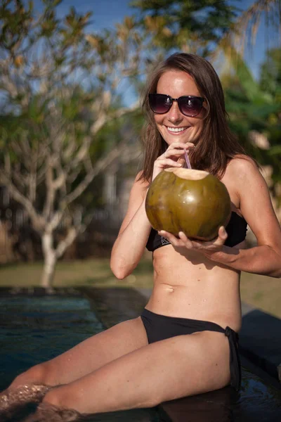 Retrato vertical de una atractiva chica sonriente en bikini negro y gafas de sol sentadas en el borde de la piscina y sosteniendo coco con paja, palmera y cielo azul en el fondo — Foto de Stock