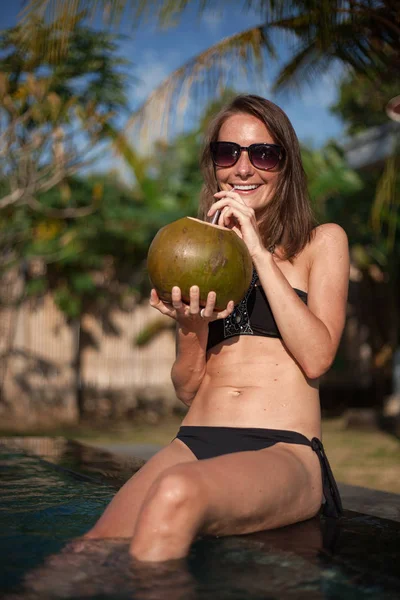 Retrato vertical de una atractiva chica sonriente en bikini negro y gafas de sol sentadas en el borde de la piscina y sosteniendo coco con paja, palmera y cielo azul en el fondo — Foto de Stock