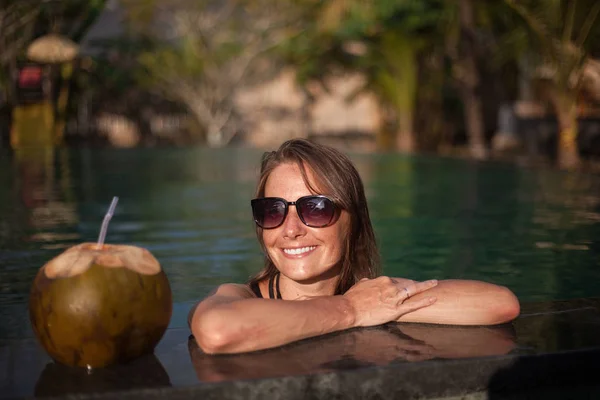 Poziome portret pięknej uśmiechający się dziewczyna w okulary siedzi w wodzie z ramionami i kokos z słomy na krawędź basenu — Zdjęcie stockowe
