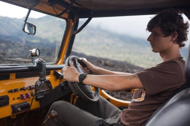 Kıvırcık saçlı dövmesi adam, Kaygılanacak bir vadi ile Bali, Endonezya volkanik kayalar üstündeki Park offroad ortalama araç içinde oturuyor ve bir ayna aracılığıyla arıyor