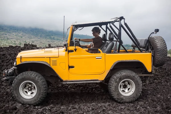 A curly-haired man is sitting in the offroad yelow vehicle turned to the right and parked at top of a valley with volcanic rock and mountains.