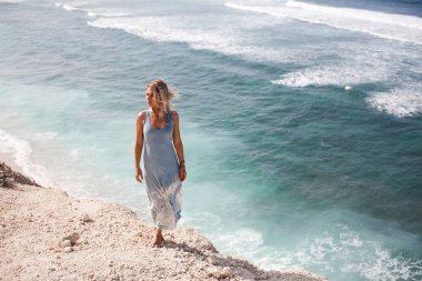 Hava ve ışık-mavi elbise içinde yüzen saçlı güzel kız rock çıkıntıya okyanus önünde duran ve uzağa arıyorsunuz