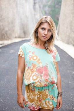 Dikey çiçekler ve soyut Yazdır ve tüy küpe ile elbiseli çekici bir sarışın kız portresi dağlar arasındaki yolda duruyor