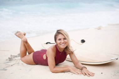 Gülümseyen güzel koyu kırmızı mayo cross-legged sörf kız bir sörf tahtası yanındaki sahilde yalan ve kamera arıyorum