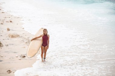 Koyu kırmızı mayo güzel sörf kız bir surfboard ile sahilde duran ve çerçevenin sol tarafında bulunan, fotoğraf arıyor