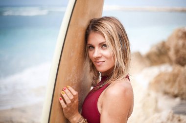 Sahilde bir surfboard holding ve kamera seyir koyu kırmızı Yüzme elbiseli çekici bir gülümseyen sörf kız yakın çekim yatay portresi