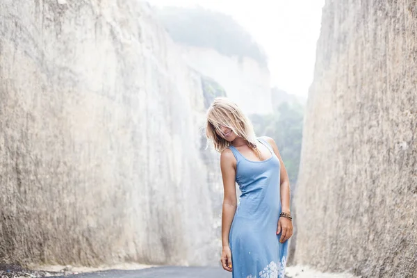 Горизонтальный портрет привлекательной улыбающейся девушки в голубом платье стоит на дороге между горами и смотрит в сторону — стоковое фото