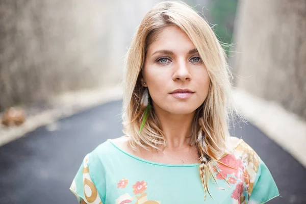 Bir çekici sarışın kız elbise soyut baskı ve tüy küpe ile gri gözleri ile yatay yakın çekim portre dağlar arasındaki yolda duruyor