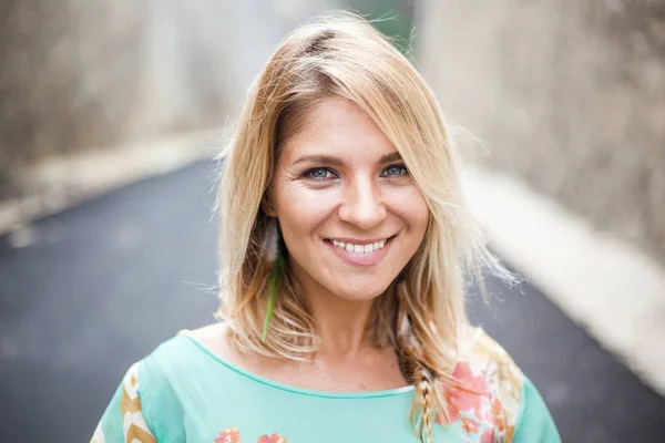 Horisontella närbild porträtt av leende blond flicka med grå ögon i klänning med abstrakt print och fjäder örhängen står på vägen mellan bergen — Stockfoto