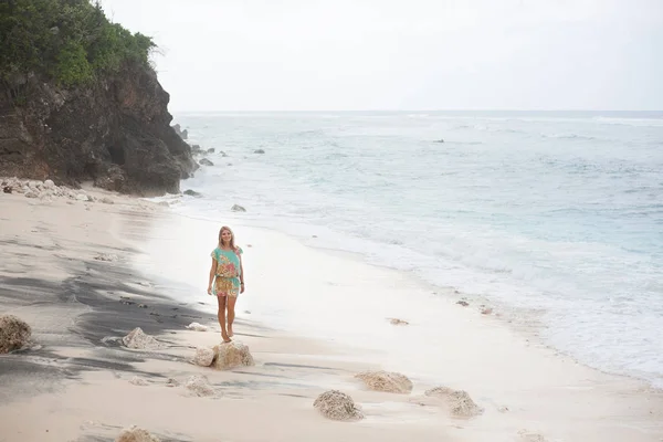 Hermosa chica rubia en vestido colorido está caminando en la playa, ubicada en el lado izquierdo del marco — Foto de Stock