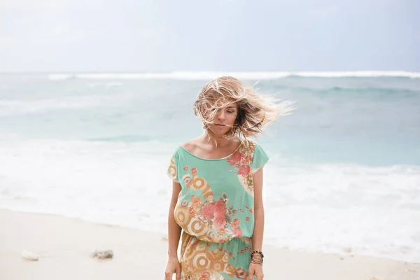 Horizontal portrait of blonde girl in colorful dress with hair floating in the air is standing near the ocean and looking down — Stock Photo, Image