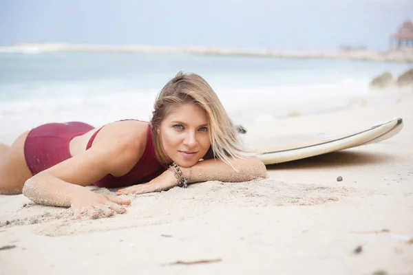 Hermosa chica de surf sonriente en traje de baño de color rojo oscuro se encuentra en la playa junto a una tabla de surf y mirando a la cámara — Foto de Stock