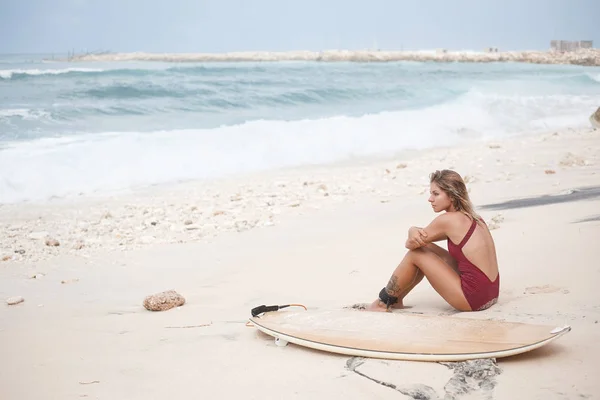 Atraktivní surfování dívka v plavkách tmavě červená sedí na pláži blízko Surf oceánu a hledat dál — Stock fotografie