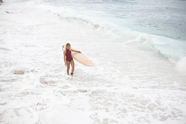 Krásná usměvavá surfování dívka v plavkách tmavě červená je chůze po pláži s surfovací prkno a hledat dál — Stock fotografie