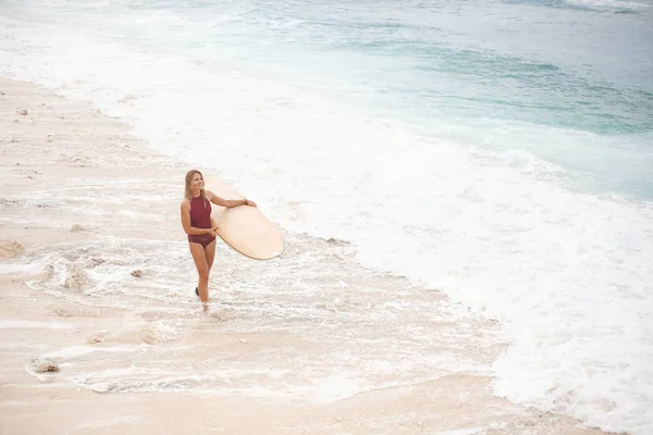 Schönes lächelndes Surfmädchen in dunkelrotem Badeanzug läuft mit einem Surfbrett am Strand entlang und schaut weg — Stockfoto