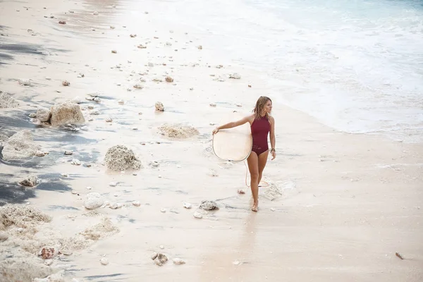 Hermosa chica de surf en traje de baño de color rojo oscuro está caminando por la playa con una tabla de surf y mirando hacia otro lado, situado en el lado derecho del marco — Foto de Stock