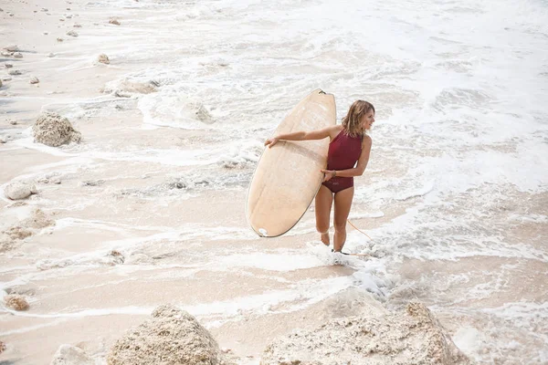 Krásný surfování dívka v plavkách tmavě červená je chůze na pláži s surfovací prkno a daleko, nachází na pravé straně rámu — Stock fotografie