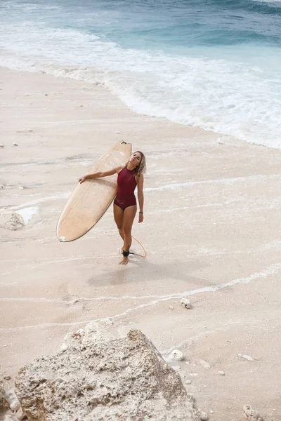 Vertikální portrét krásné surf Girl v tmavě červených plavkách s surfovací prkno se děje z oceánu a zavřel oči — Stock fotografie