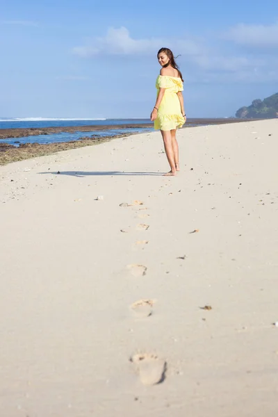 Retrato vertical de la hermosa chica sonriente en vestido amarillo alejándose a lo largo de la playa y sus huellas — Foto de Stock