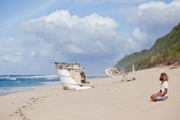 Robinson Crusoe. Curly-bebaarde man zit op het strand met een schip schipbreuk op de backgraund. — Stockfoto
