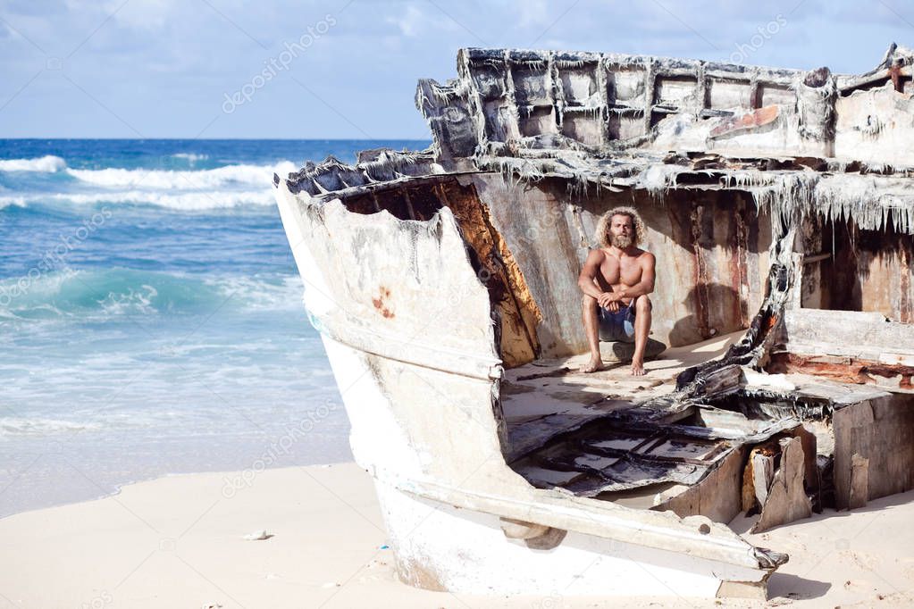 Robinson Crusoe. Curly-bearded man is sitting on the wreck of the ship on the beach